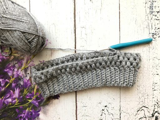 A grey crochet piece with a crochet hook and purple flowers on a wooden surface.