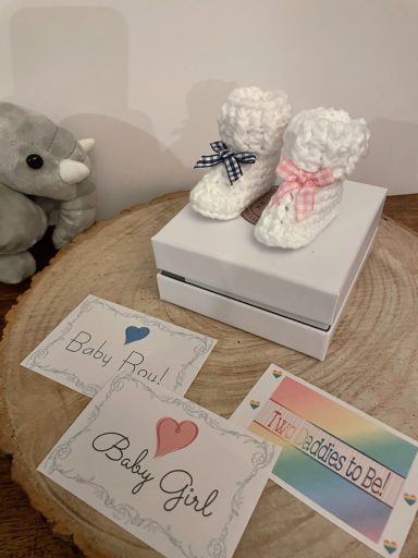 White baby booties with pink and blue bows, alongside "Baby Girl" cards on a wooden table.